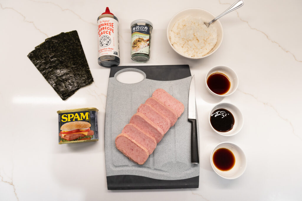 The ingredients for spam musubi laid out: a spam block cut into six slices, a bowl of cooked rice, sheets of nori, a bottle of furikake, and three bowls with soy sauce, teriyaki sauce, and oyster sauce.