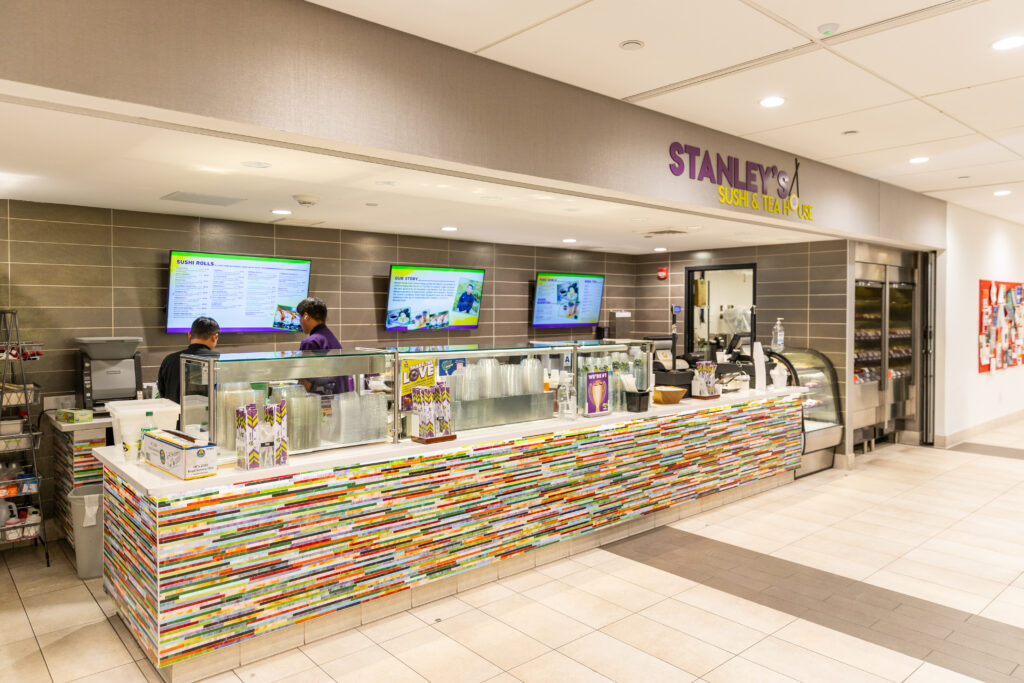 Stanley's Sushi ordering counter with a colorful tile mosaic and menu TV screens on the wall behind.